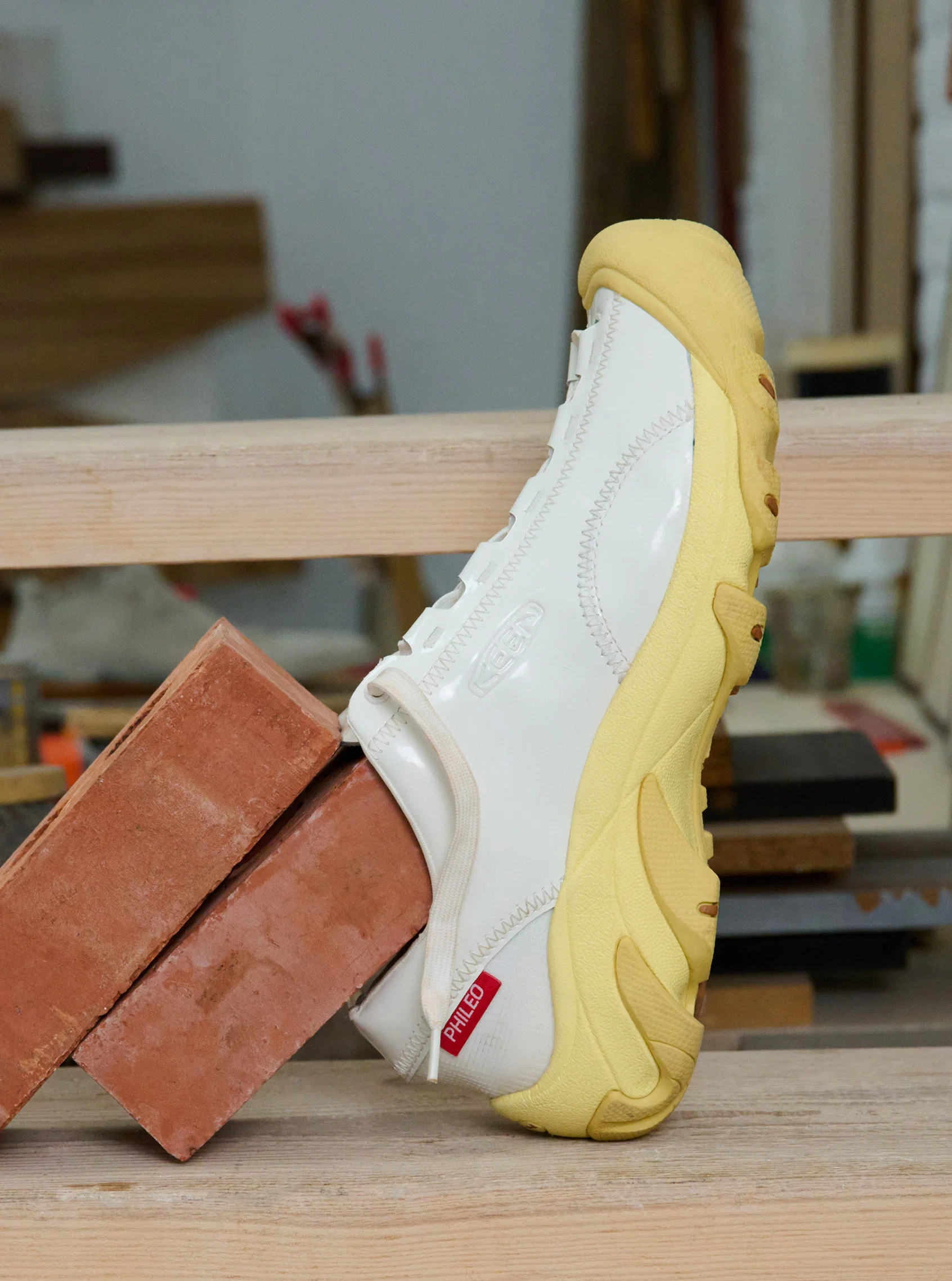 White and yellow Phileo collaboration Targher sneaker leaned up against bricks in a woodworking shop.
